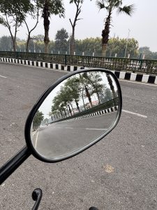 A close-up view of a motorcycle's side mirror reflecting a deserted road lined with trees and a green fence. The road is largely empty, with a faint silhouette of a motorcycle visible in the distance. 